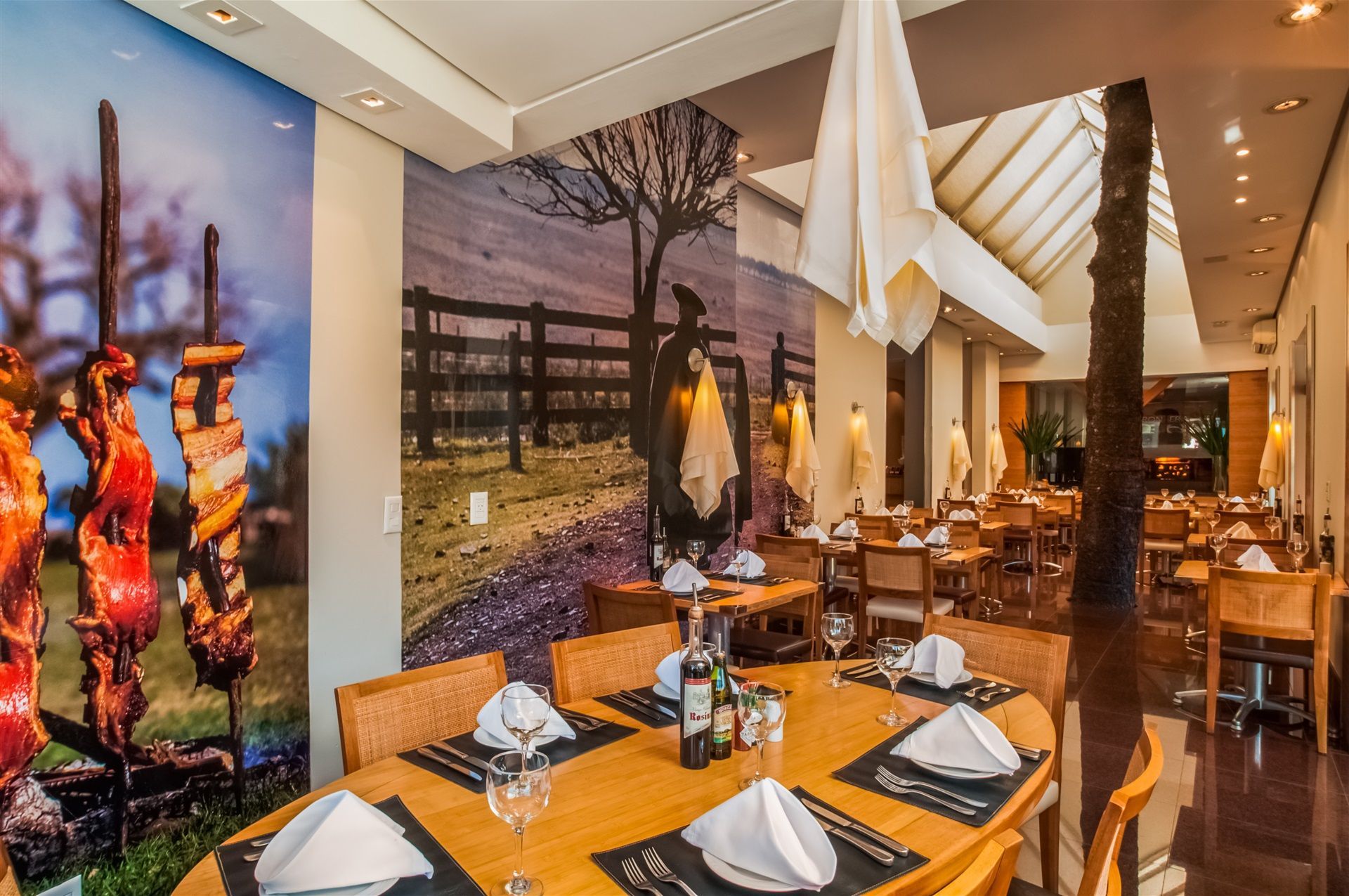 Sala de jantar elegante do restaurante com mesas postas, iluminação aconchegante e grandes murais nas paredes com cenas de vinho e vinhedos.