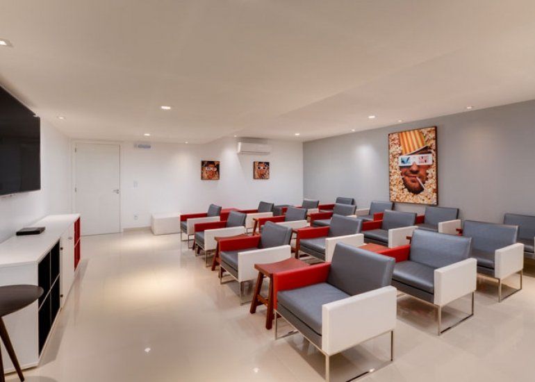 Sala de espera moderna com cadeiras cinza e vermelhas, paredes brancas, iluminação embutida e obras de arte nas paredes.