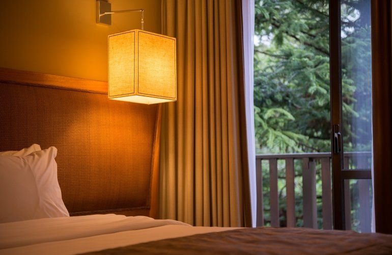 Quarto aconchegante com luz quente de abajur, cama e varanda com vista para as árvores através de portas de vidro deslizantes.