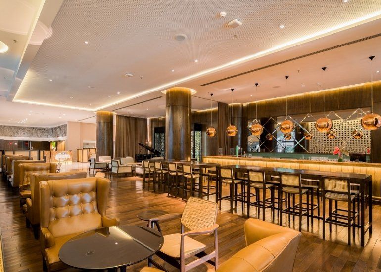 Lounge bar moderno com cadeiras estofadas, banquetas altas e iluminação aconchegante em um interior elegante.