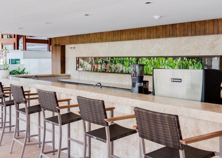 Bar de hotel moderno com banquetas, balcão longo de mármore e máquina de café sob uma parede revestida de madeira.