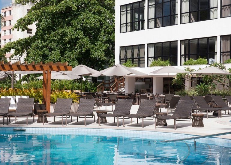 Área da piscina do resort com espreguiçadeiras, guarda-sóis e um prédio de hotel moderno ao fundo.