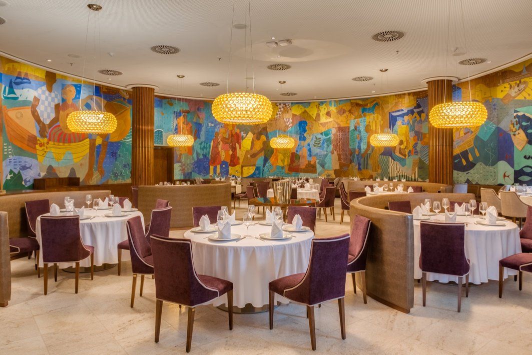 Sala de jantar elegante de restaurante com cadeiras roxas, mesas com toalhas brancas e paredes com murais coloridos.