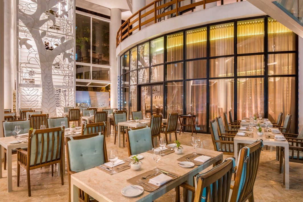 Sala de jantar elegante de restaurante com mesas postas, cadeiras listradas e grandes janelas em arco.