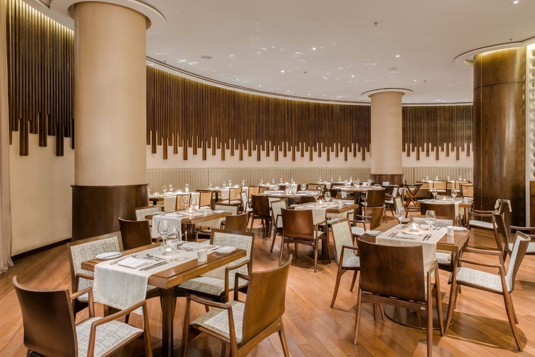 Interior de restaurante moderno e elegante com mesas postas, cadeiras em tons de bege e detalhes em madeira aconchegantes.