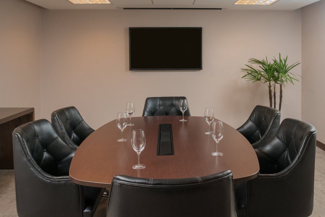 Sala de conferências com mesa oval, cadeiras de couro preto, TV fixada na parede e planta em vaso.