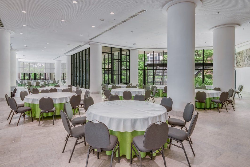 Salão de banquetes luminoso com mesas redondas, cadeiras cinzentas, toalhas de mesa brancas e grandes janelas com vista para a vegetação.