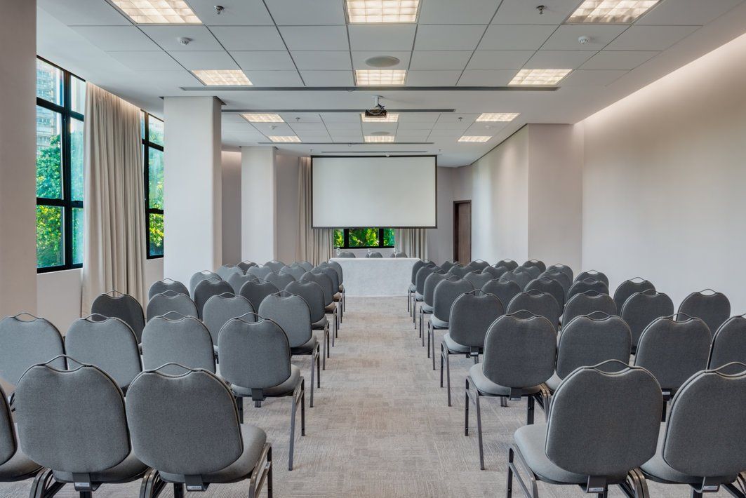 Sala de conferências vazia com fileiras de cadeiras cinzentas voltadas para uma tela de projeção e grandes janelas.
