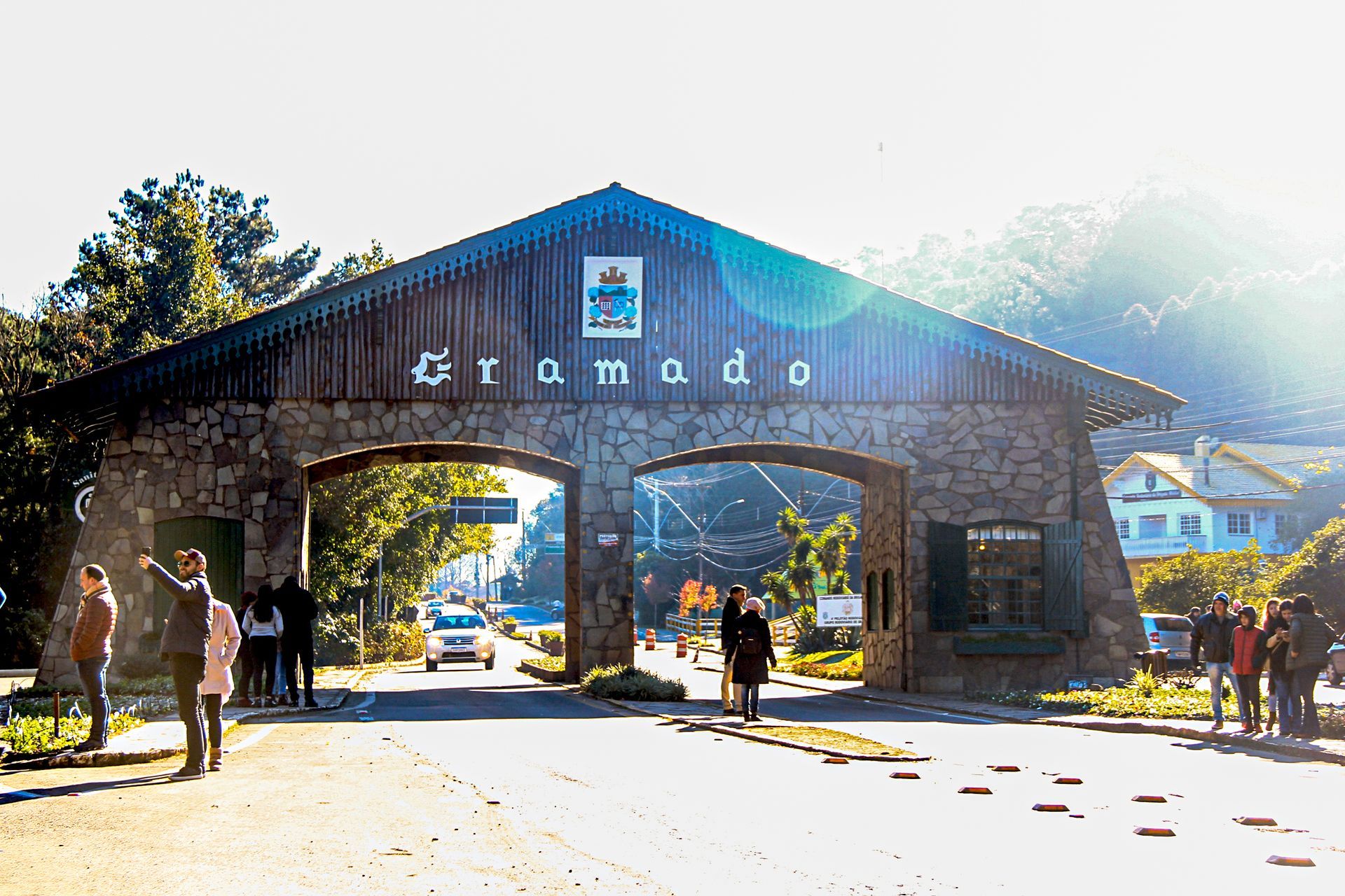 Pessoas caminhando sob um grande arco de pedra em um portão de entrada ensolarado com montanhas ao fundo.