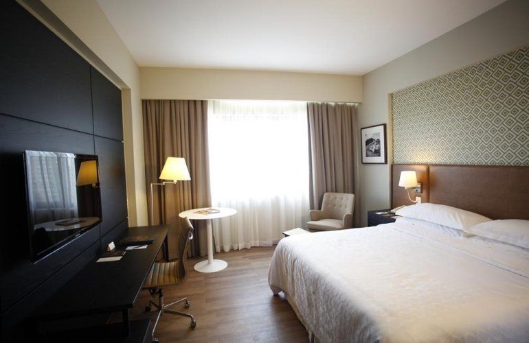 Quarto de hotel com cama king-size, cadeira, abajur, TV e cortinas claras na janela.