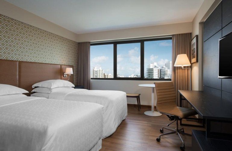 Quarto de hotel com duas camas, escrivaninha, TV e janela ampla com vista para os prédios da cidade.
