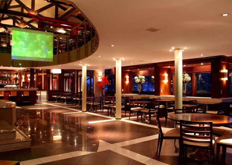 Interior vazio de um restaurante sofisticado com pisos polidos, mesas, cadeiras e uma grande tela de TV verde.