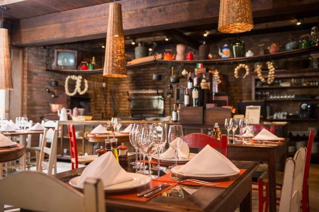 Sala de jantar aconchegante do restaurante, com mesas postas, taças de vinho, guardanapos e prateleiras rústicas de madeira atrás do bar.