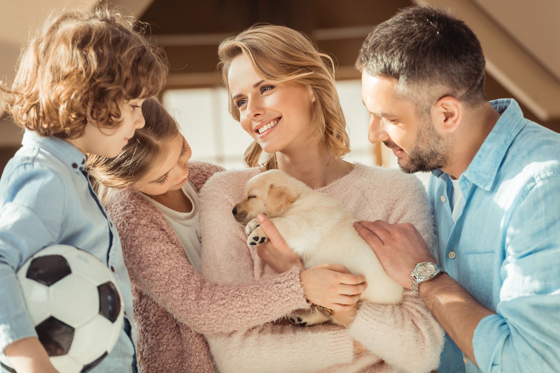 Família abraçando um cachorro branco e fofo ao lado de uma bola de futebol, sorrindo em um ambiente externo ensolarado.