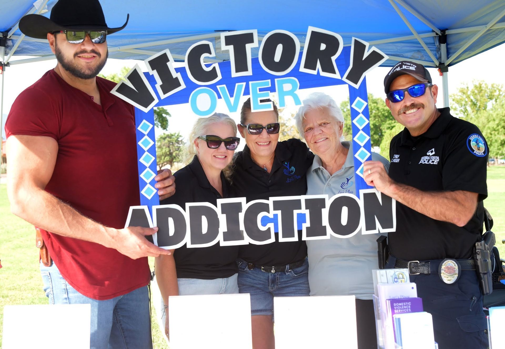 Four people smiling under a tent hold a “Victory Over Addiction” sign at an outdoor event