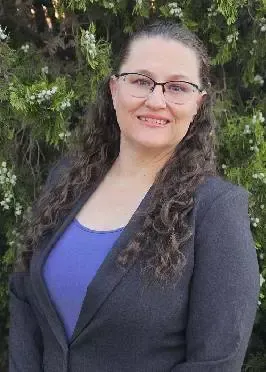 A woman wearing glasses and a gray jacket is standing in front of a tree.