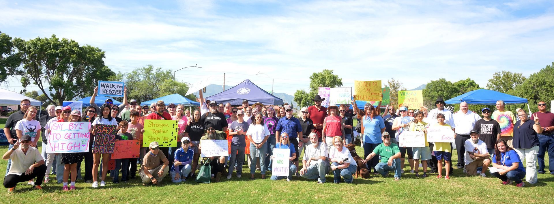 Large group posing outdoors at a community gathering, holding colorful signs under tents on a sunny day