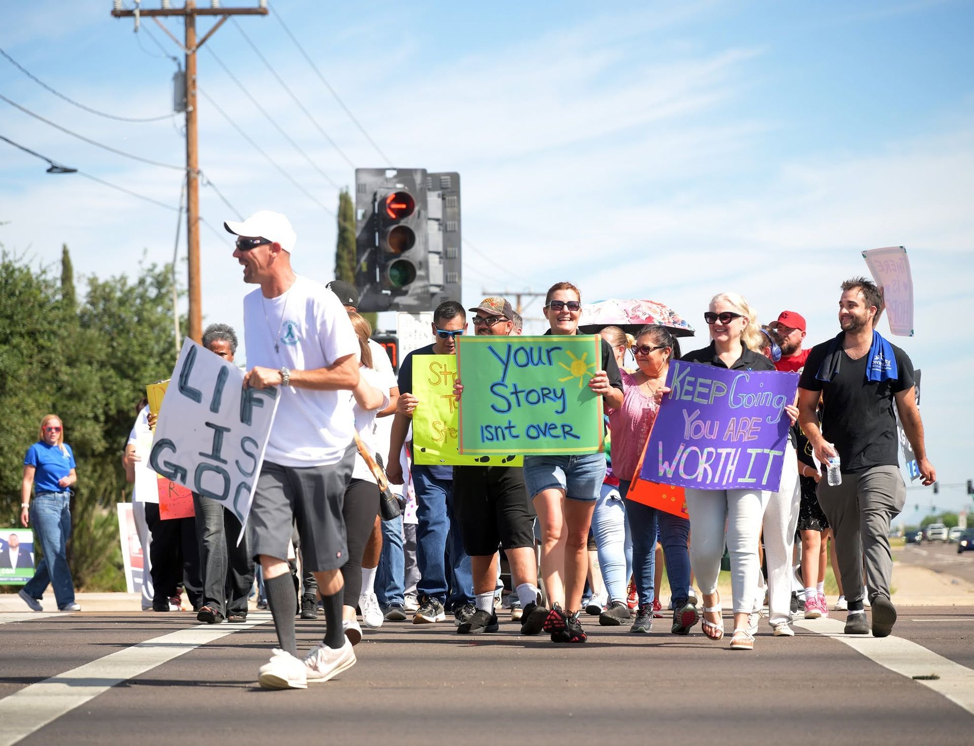 People holding colorful protest signs march across a street at a crosswalk under a traffic light.