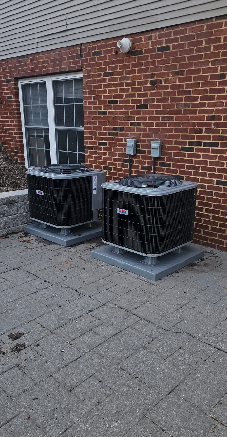 Two black HVAC units on concrete pads beside a brick building wall.