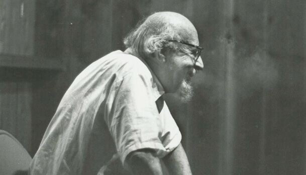 Una persona con gli occhiali e la barba espira una nuvola di fumo, ripresa di profilo contro una parete di legno.