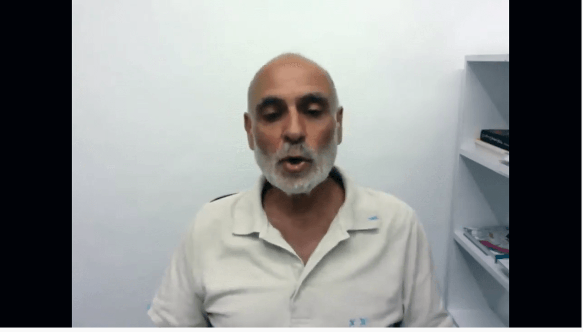 Una persona con la barba, che indossa una polo di colore chiaro, è in piedi davanti a una parete bianca con una libreria sulla destra.