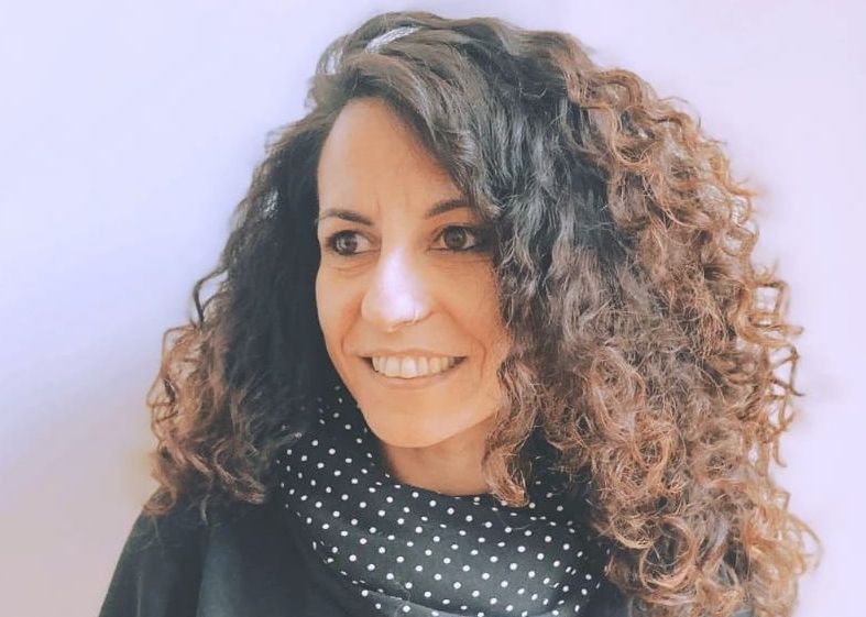 Una persona sorridente con lunghi e voluminosi capelli scuri e ricci, che indossa un abito nero a pois bianchi.