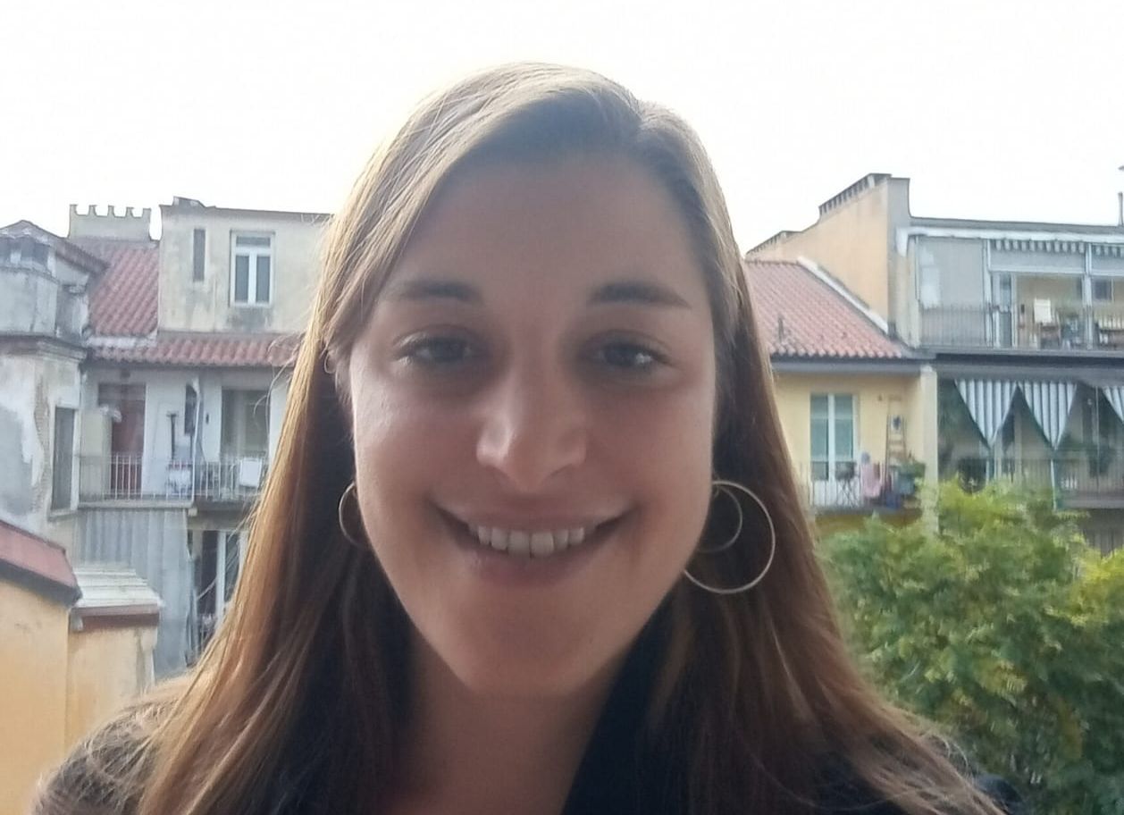 Una persona con lunghi capelli castani e orecchini a cerchio sorride, con edifici e un albero sullo sfondo sfocato.