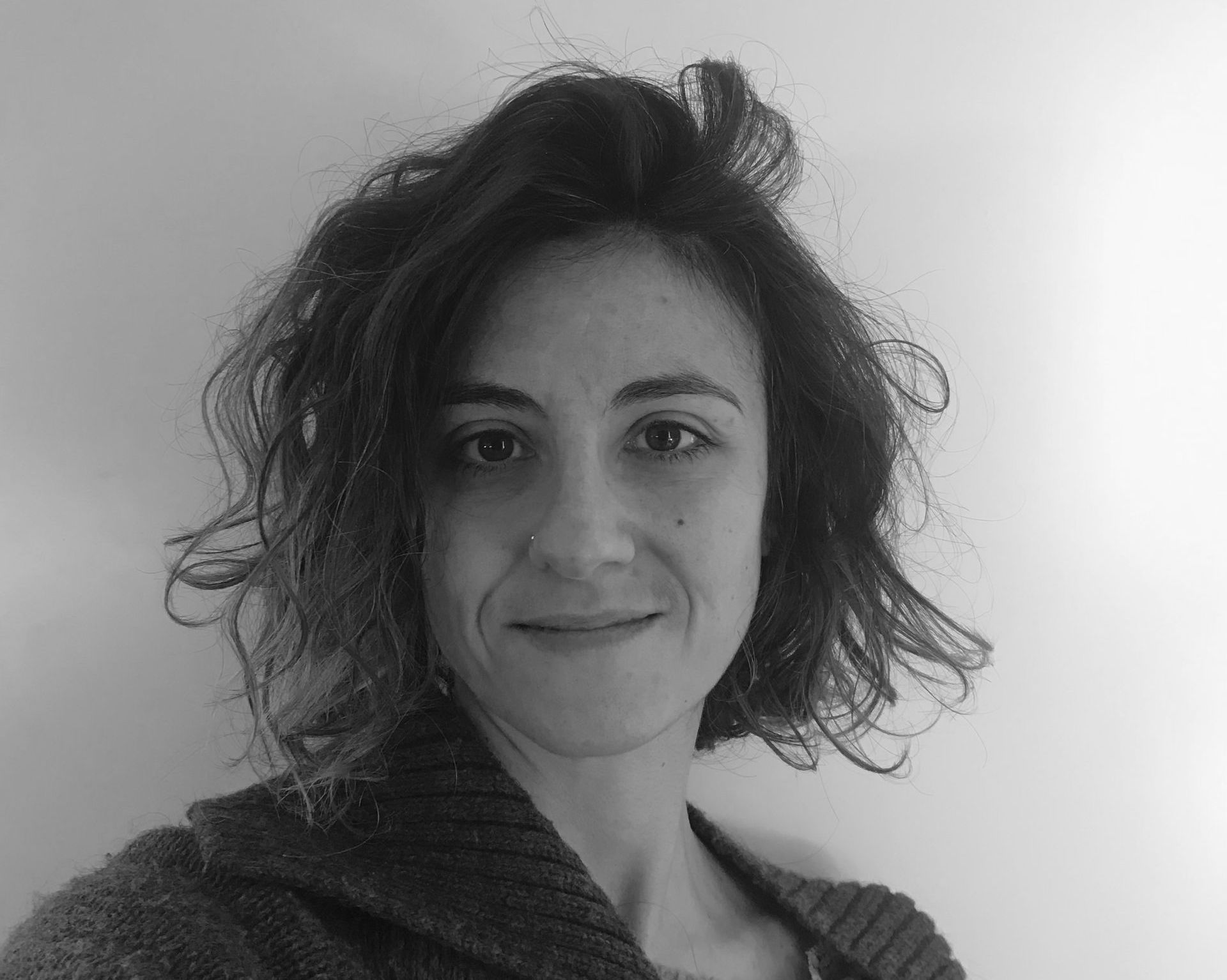 Primo piano del volto di una persona con capelli scuri e ricci, occhi chiari e una leggera barba incolta, che sorride dolcemente su uno sfondo sfocato.