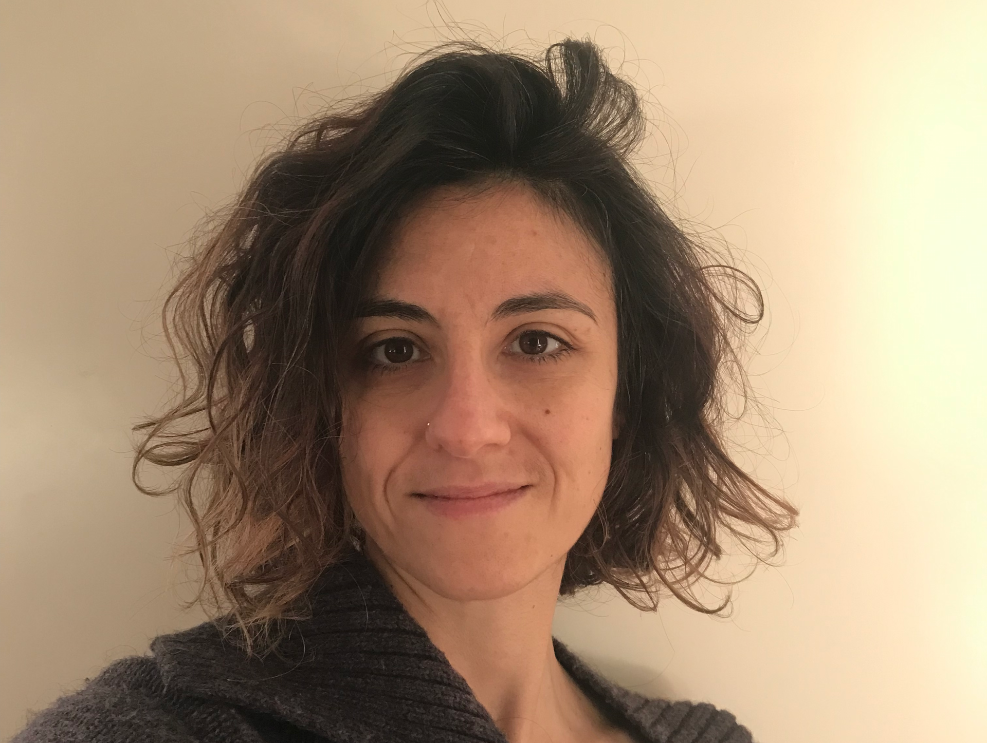 Una persona con capelli castani ondulati lunghi fino alle spalle e un piercing al naso guarda in camera, sullo sfondo di una semplice parete color bianco sporco.