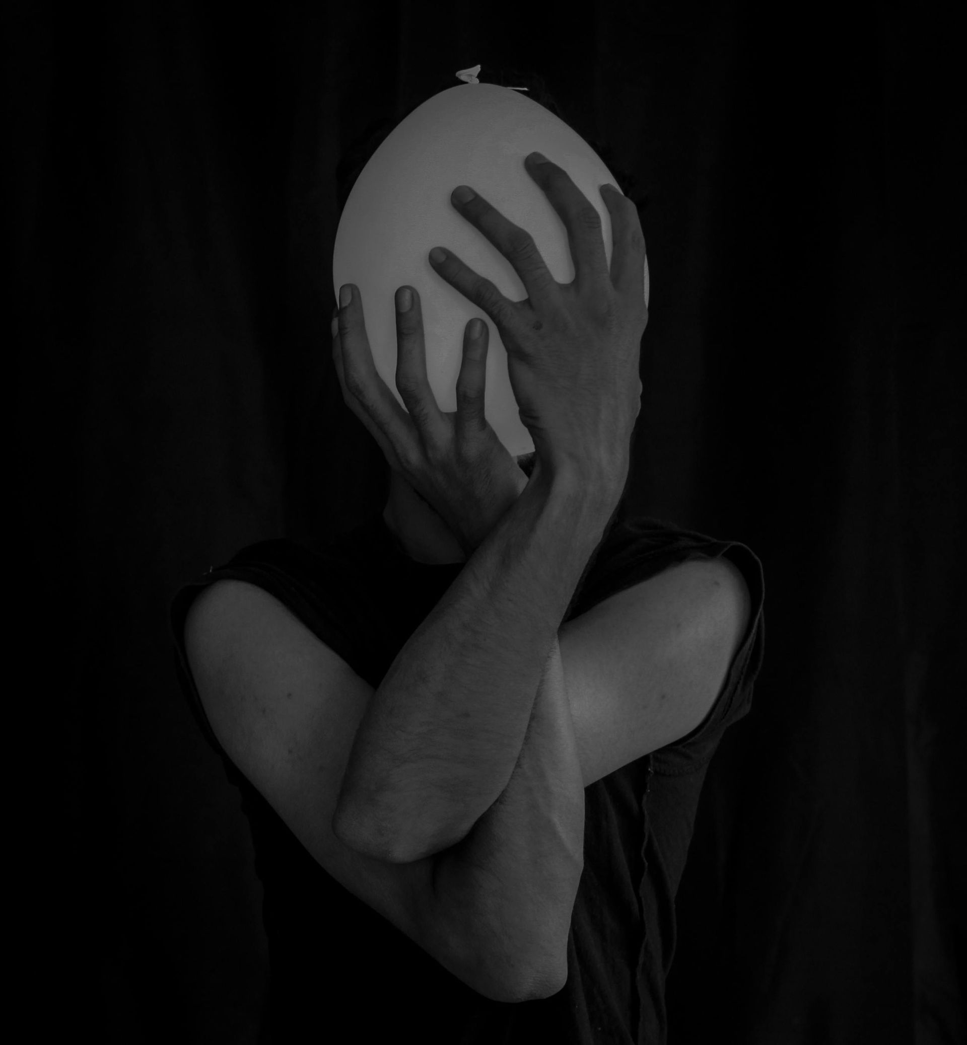 Una persona con una maglietta nera tiene un palloncino bianco davanti al viso con entrambe le mani, a braccia incrociate, su uno sfondo scuro.
