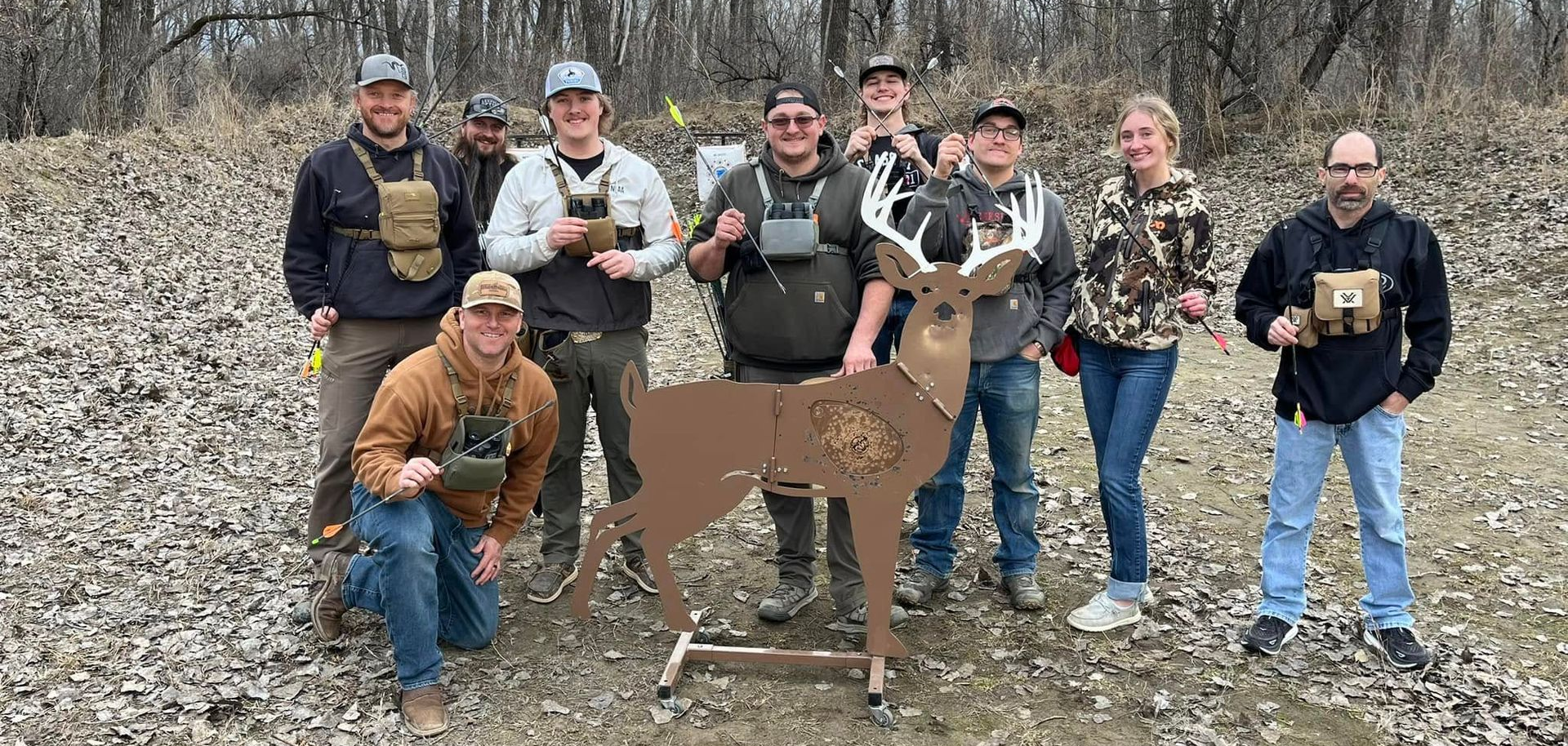 Group of people posing with a deer-shaped archery target outdoors, wearing hunting gear and smiling.