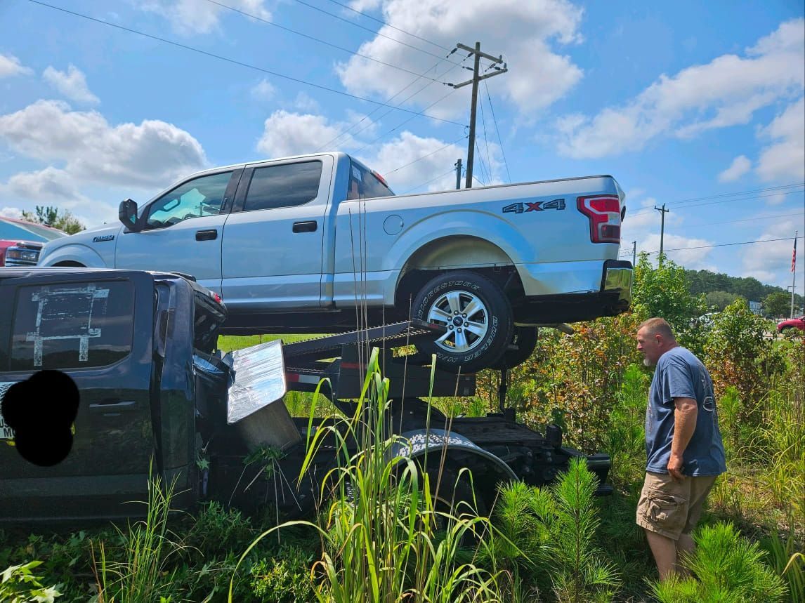 Silver pickup truck perched on a trailer, with a man standing nearby in a grassy lot under a blue sky.