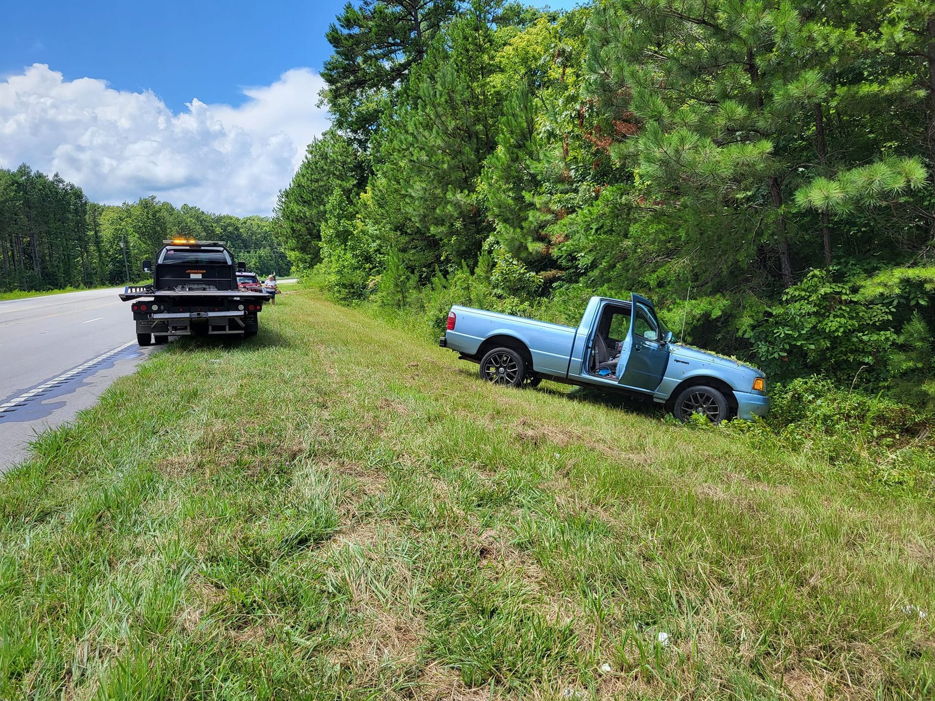 Blue pickup truck off the road in a grassy ditch beside a highway, with a tow truck nearby.