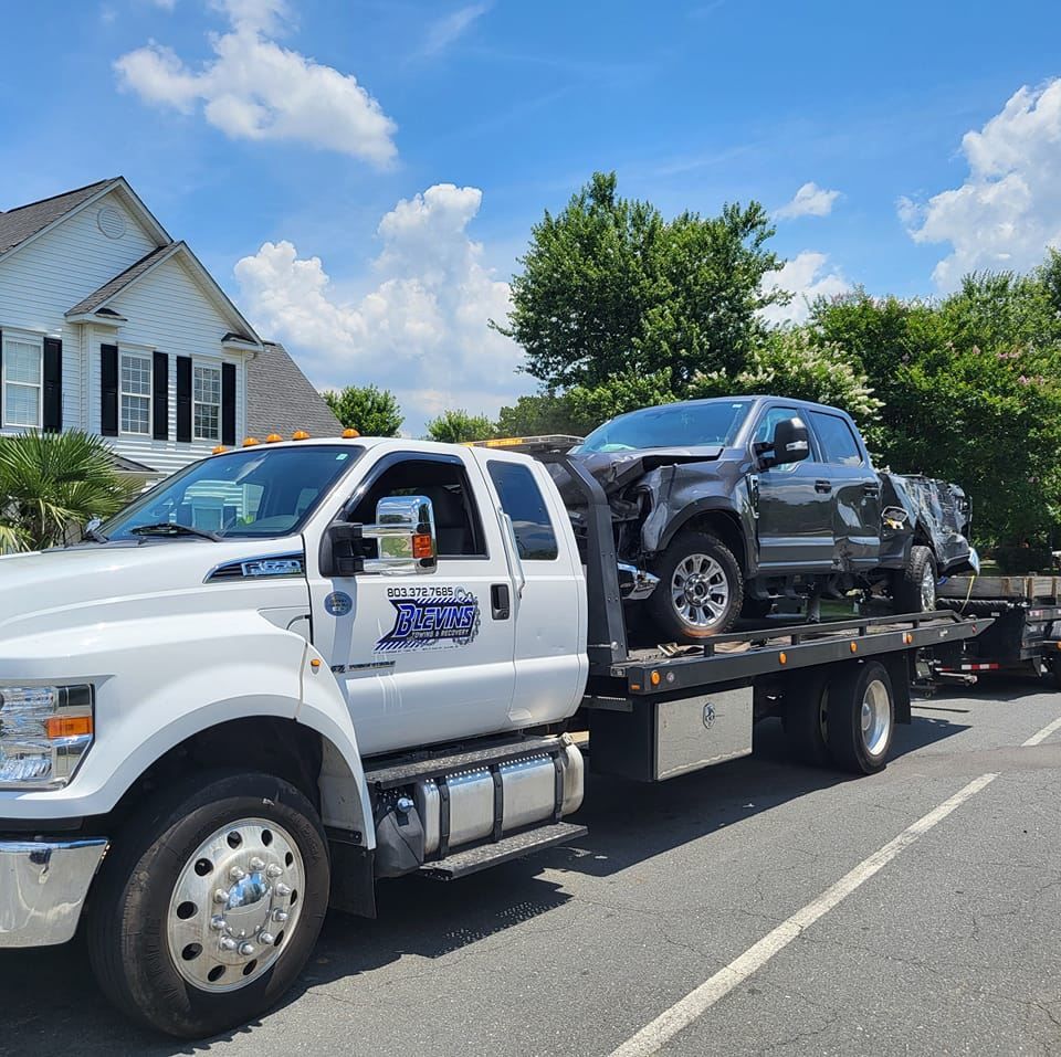 Tow truck hauling a damaged black SUV on a flatbed along a residential street under a blue sky