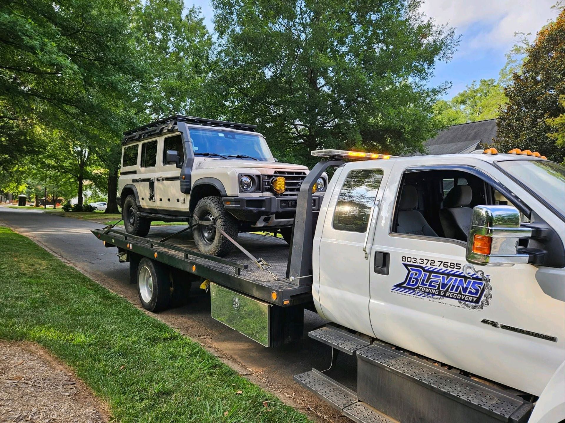 Tow truck hauling a white Jeep on a flatbed along a tree-lined street
