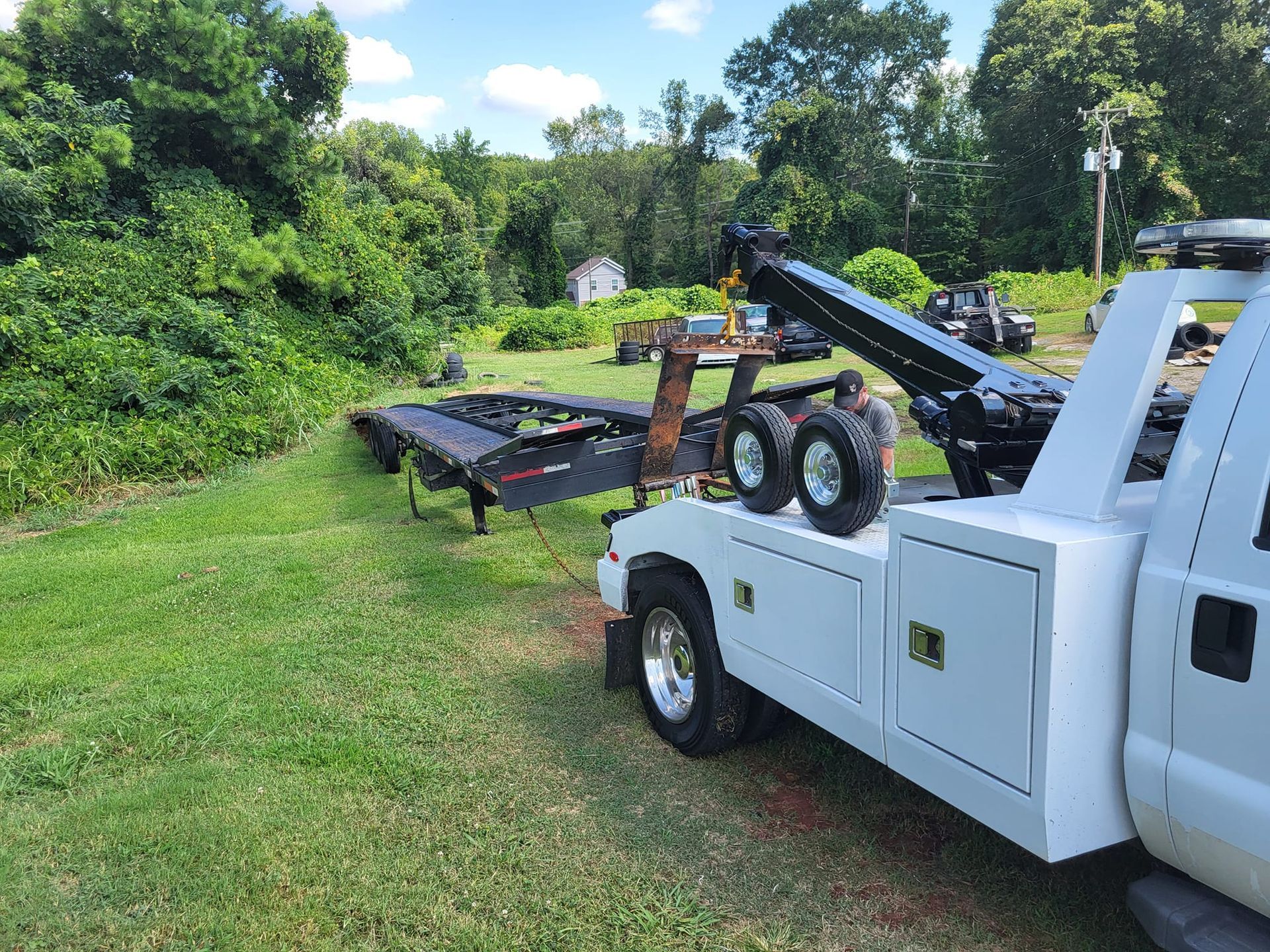 White tow truck carrying a black flatbed trailer on a grassy roadside, with trees in the background