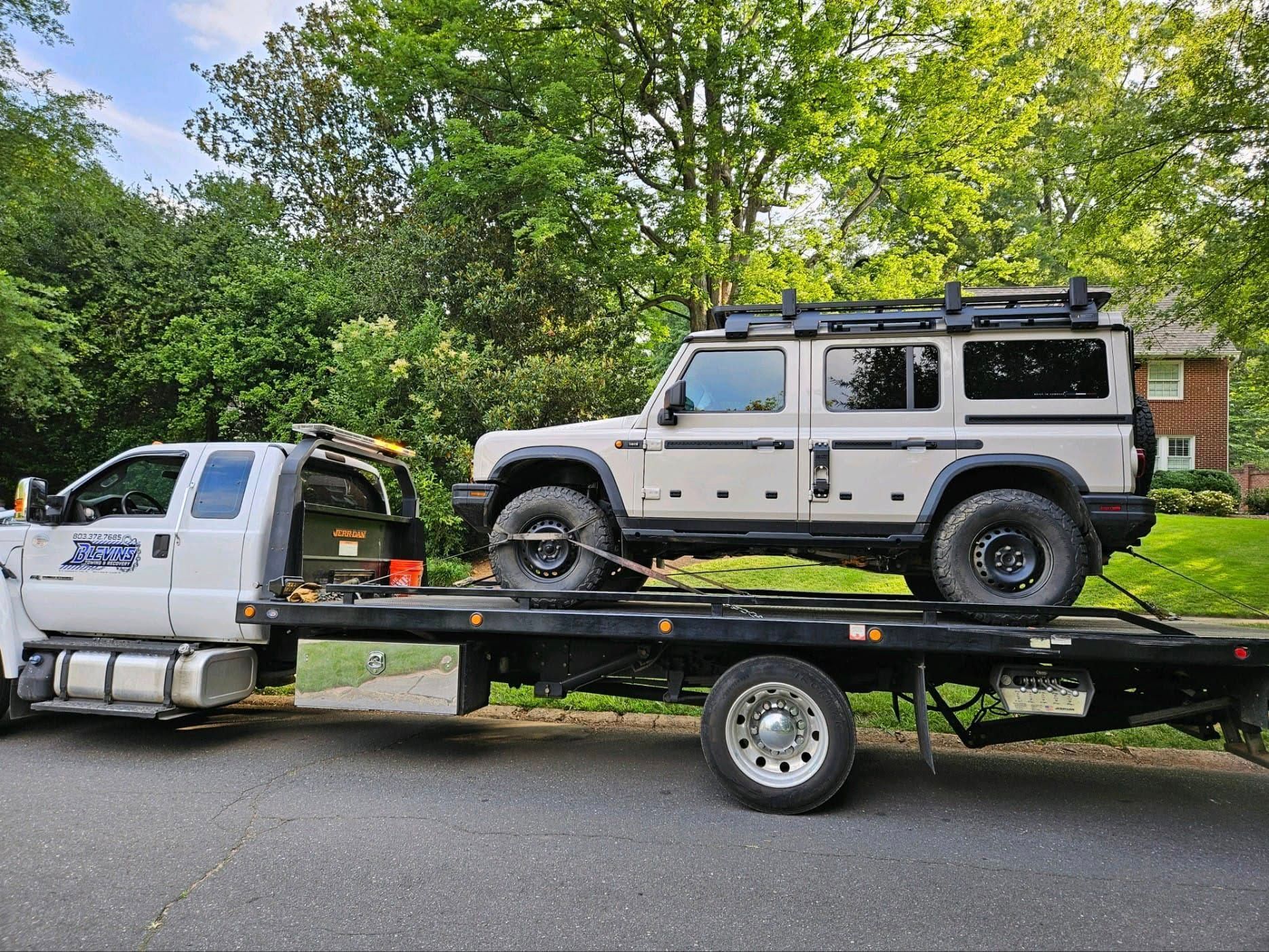White Jeep Wrangler on a flatbed trailer behind a white truck, parked outdoors under trees.