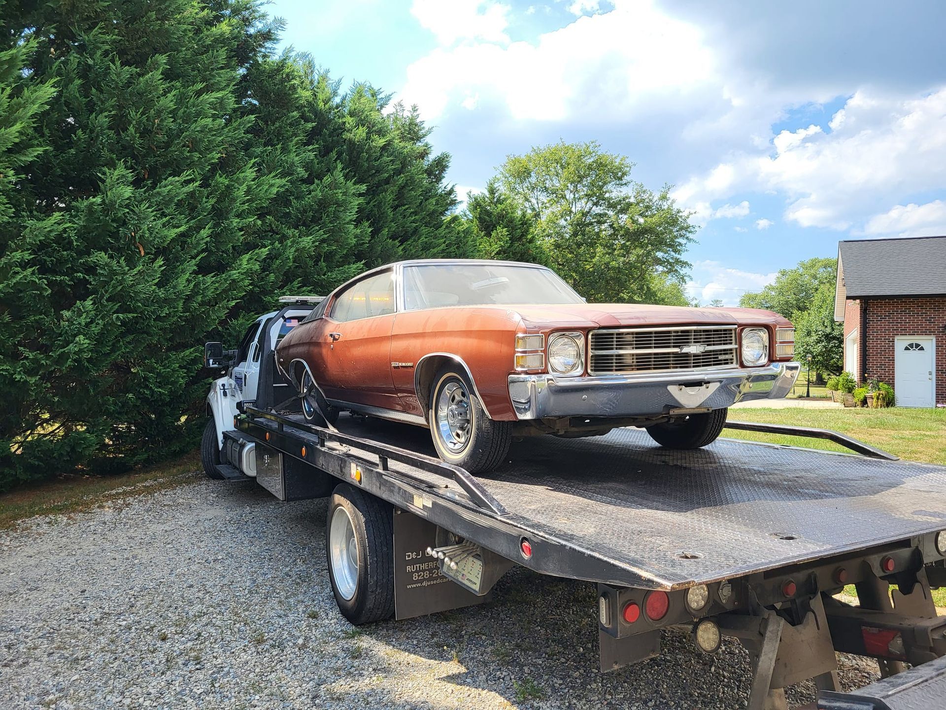 Copper classic car on a flatbed tow truck outdoors beside trees and a house