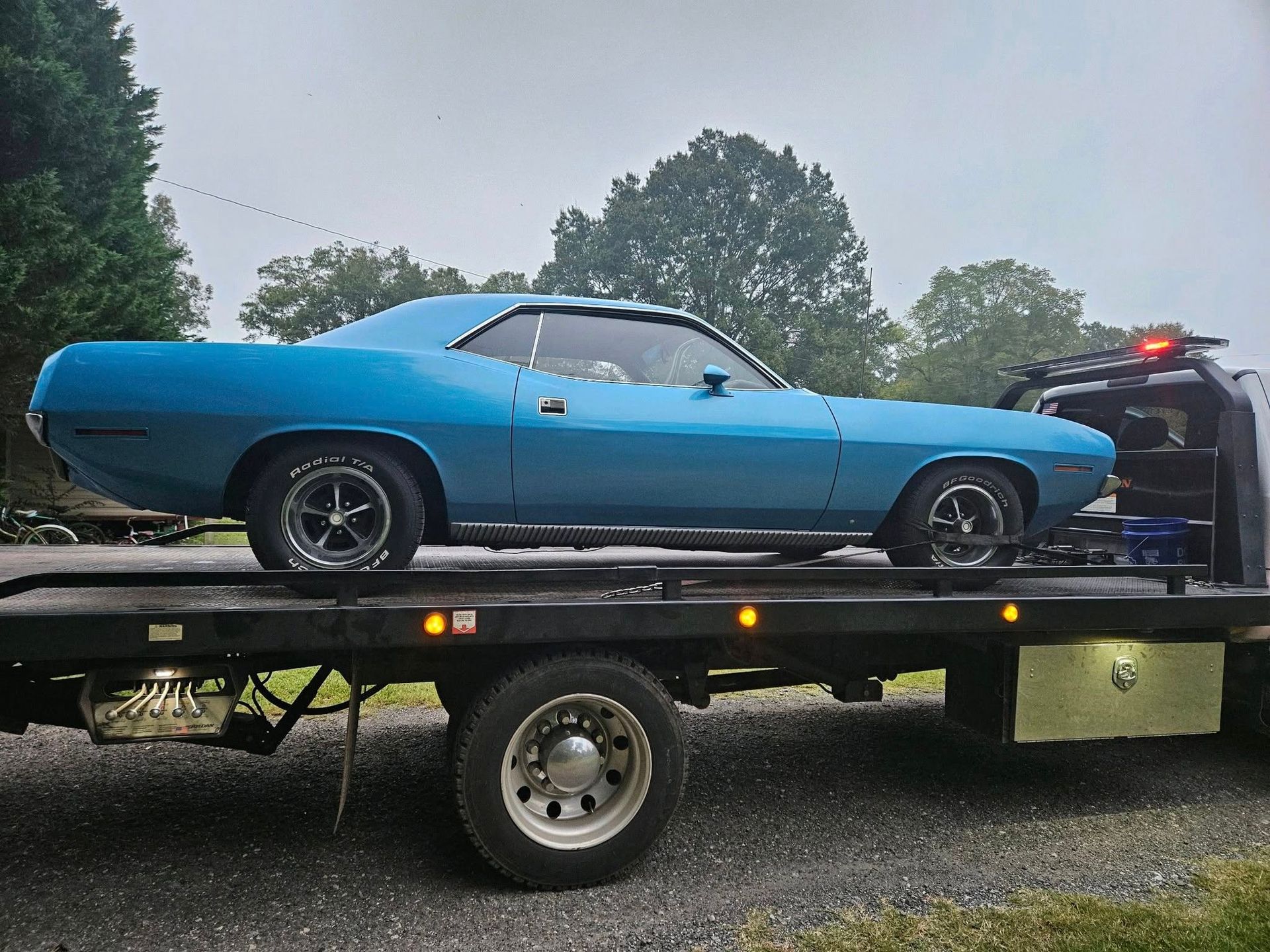 Blue classic muscle car on a flatbed trailer outdoors, with another vehicle behind it.