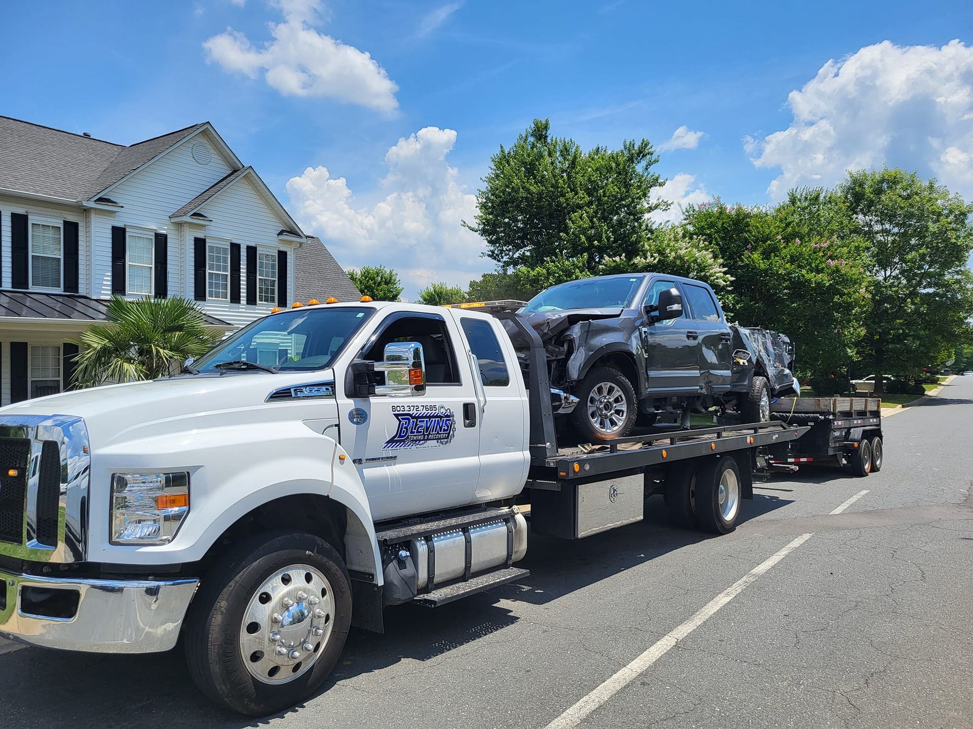 White flatbed tow truck carrying a black SUV on a sunny residential street near houses.