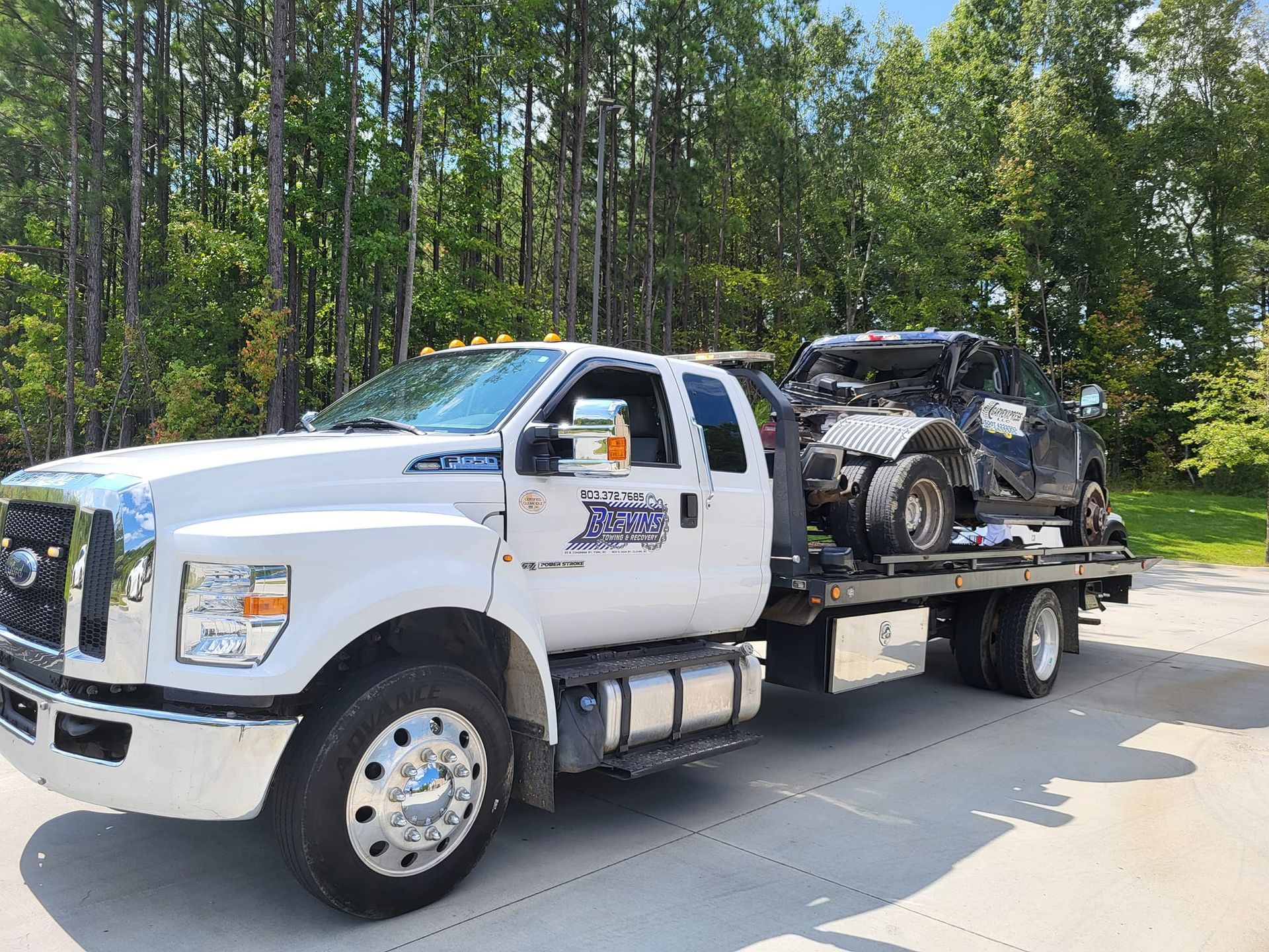 White flatbed tow truck carrying a wrecked dark vehicle on a road by trees