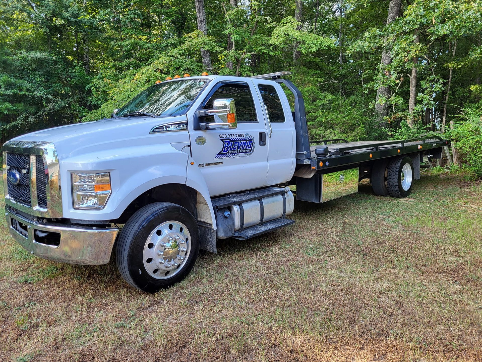 White flatbed tow truck parked on grass beside a wooded area
