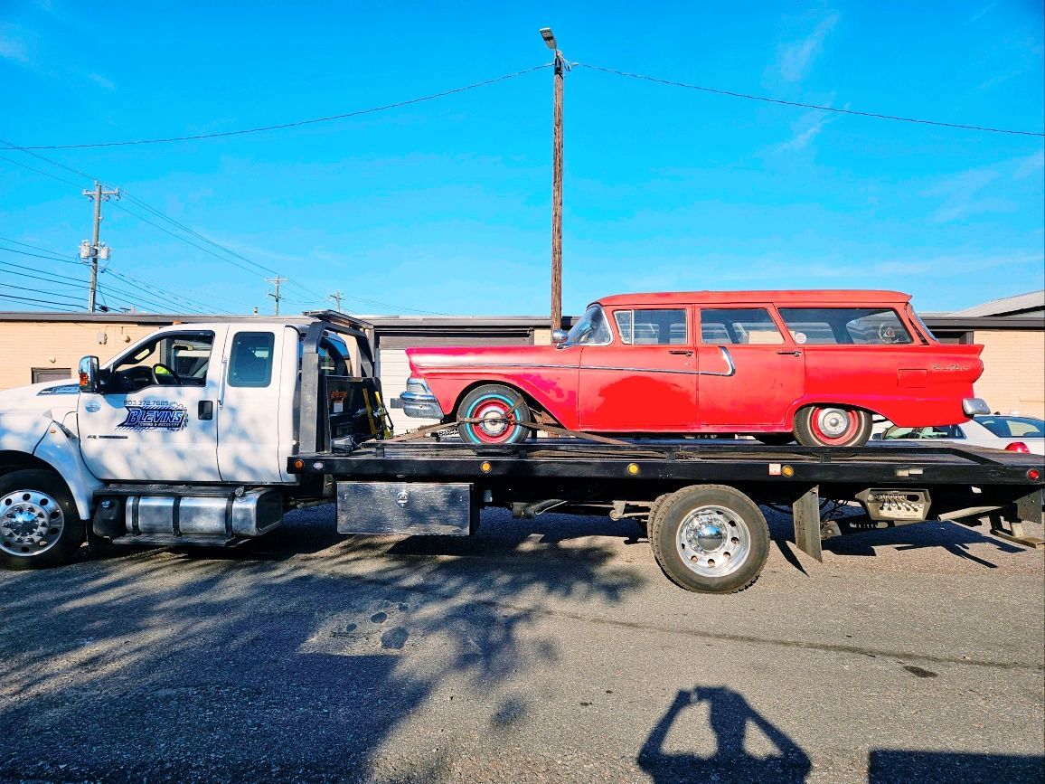 Tow truck carrying a red vintage station wagon on a flatbed in a sunny parking lot