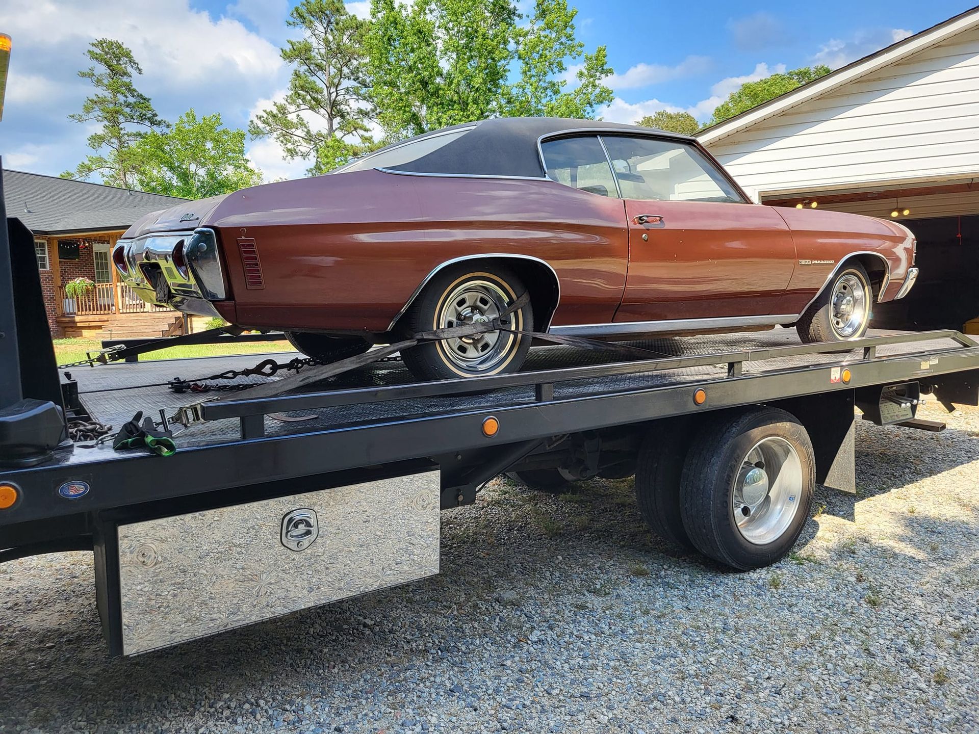 Brown classic convertible on a black car trailer in a driveway, with trees and a house in the background