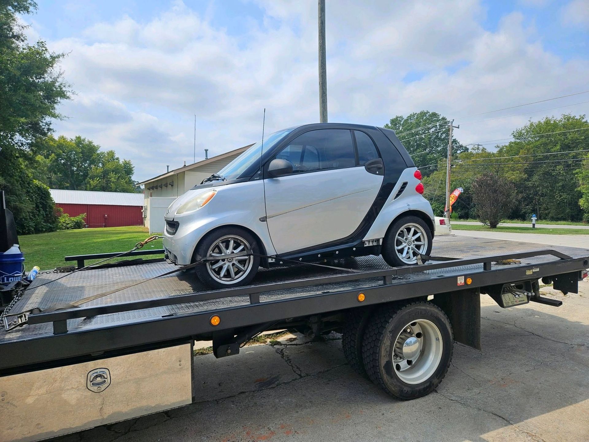 A small white and black Smart car strapped on a flatbed trailer outdoors.