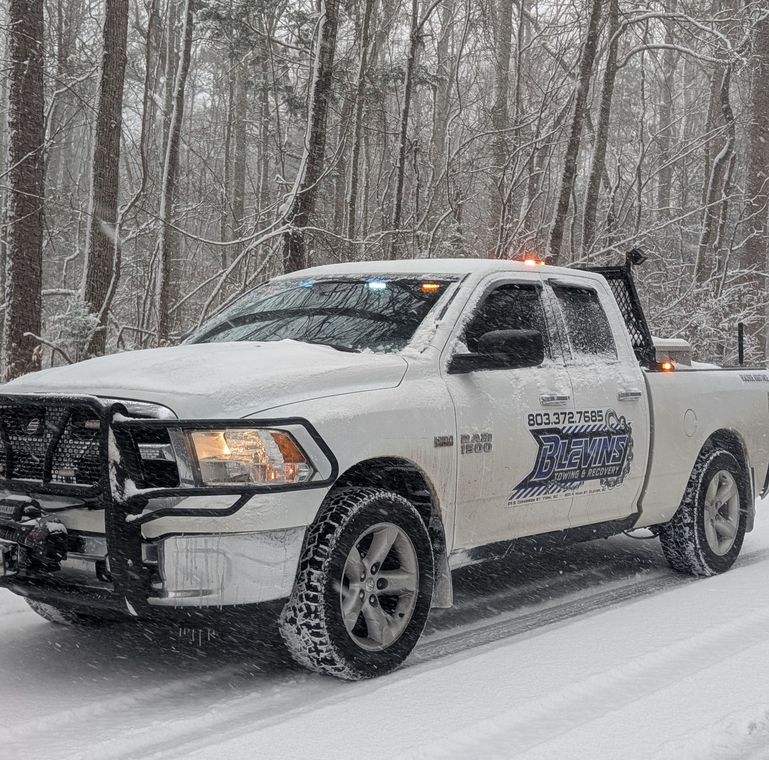 White pickup truck with a snow plow driving through a snowy forest road