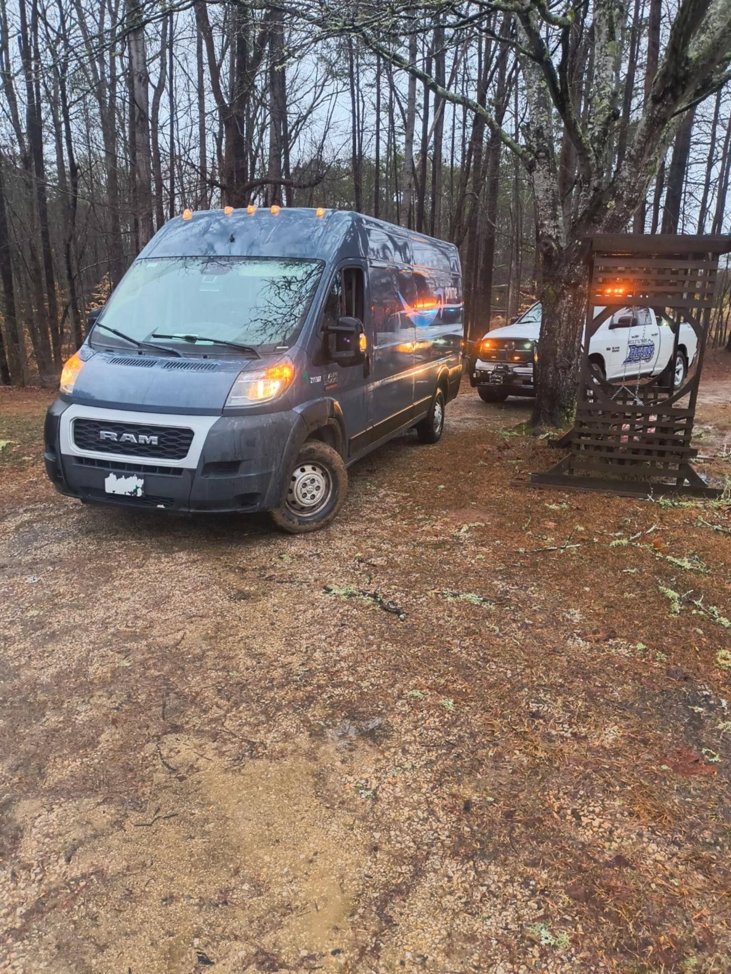 Two parked vans in a muddy wooded area, one dark blue and one white with orange lights on.
