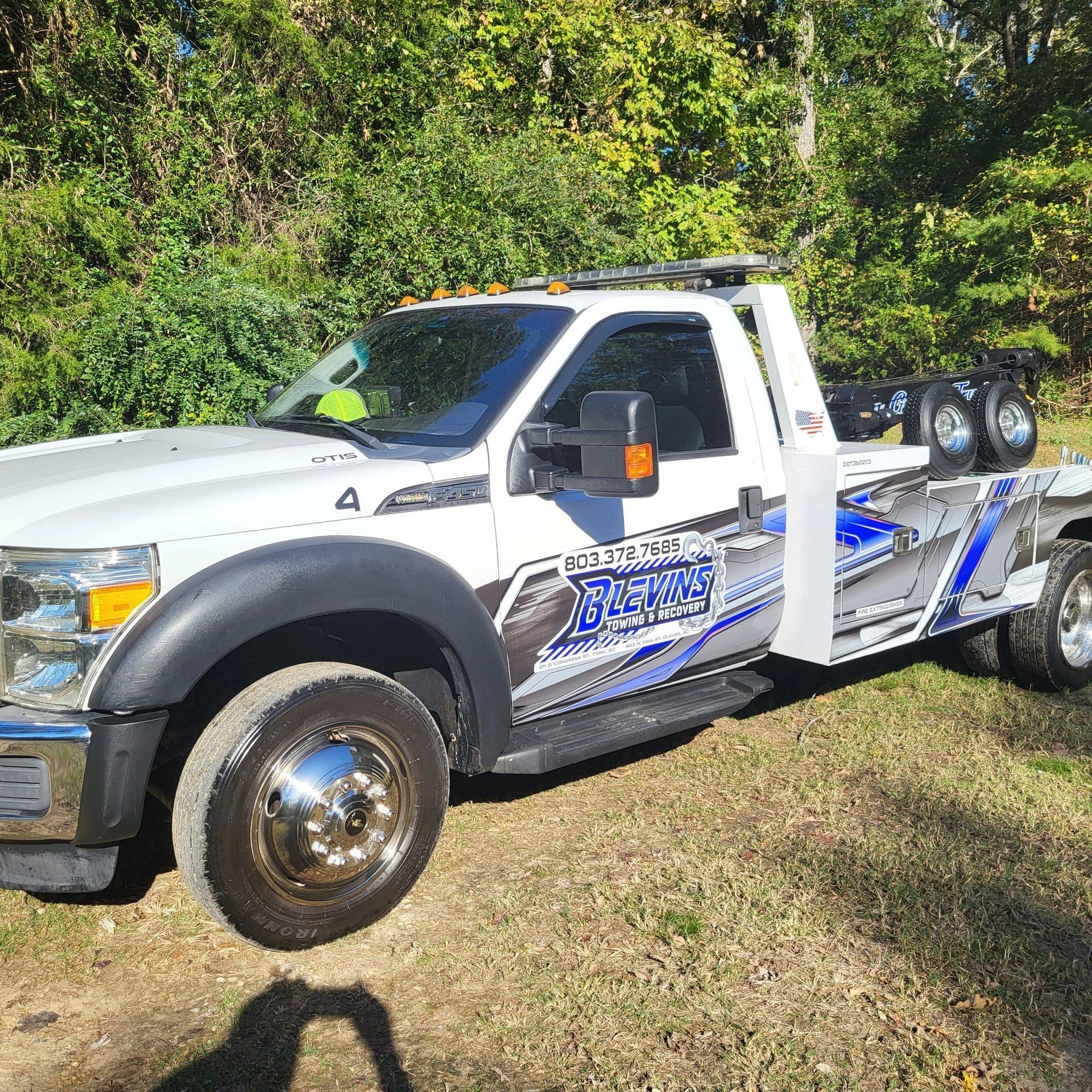 White tow truck parked on grass beside trees, with blue graphics and rear-mounted equipment