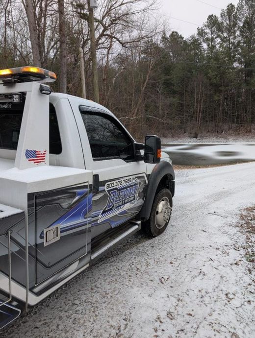 Tow truck on a snowy road beside a wooded area, with amber lights flashing.