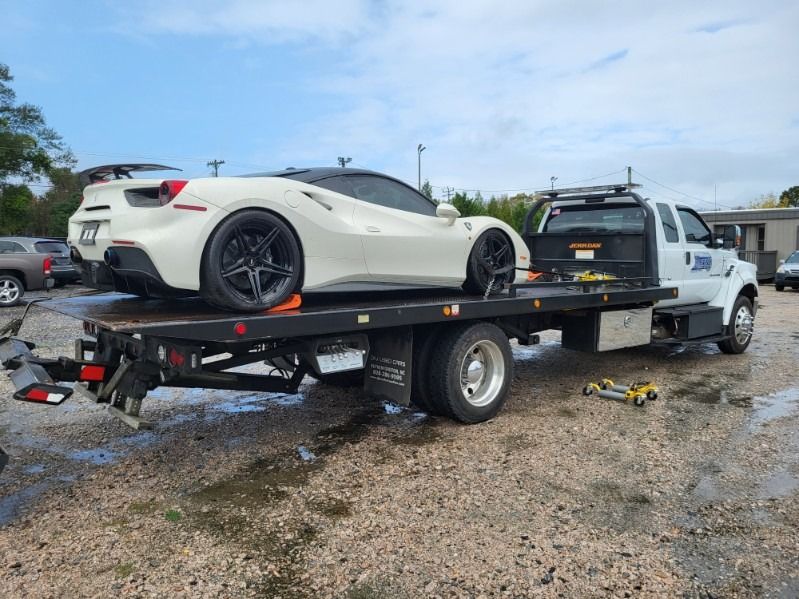 White sports car on a flatbed tow truck in a muddy lot, with a white pickup attached behind.